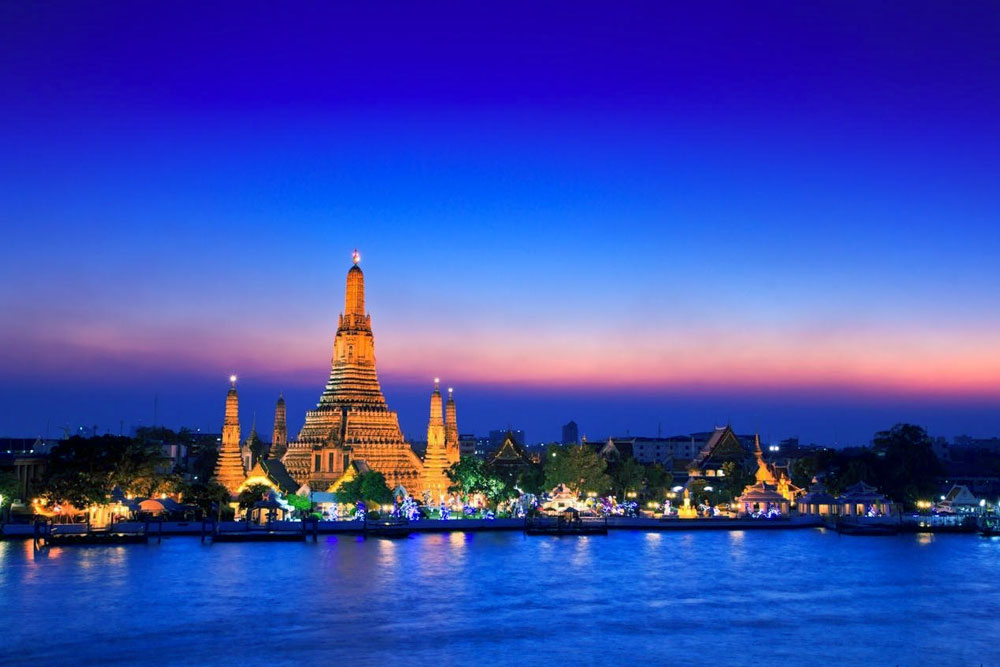 Illuminated ornate temple spire beside a river at twilight, vivid blue and orange sky, serene and scenic mood